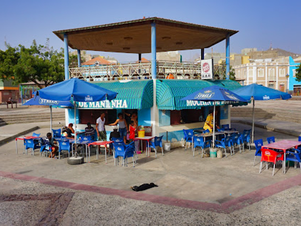 Outdoor food kiosk with blue awnings and tables in a paved square in Mindelo.