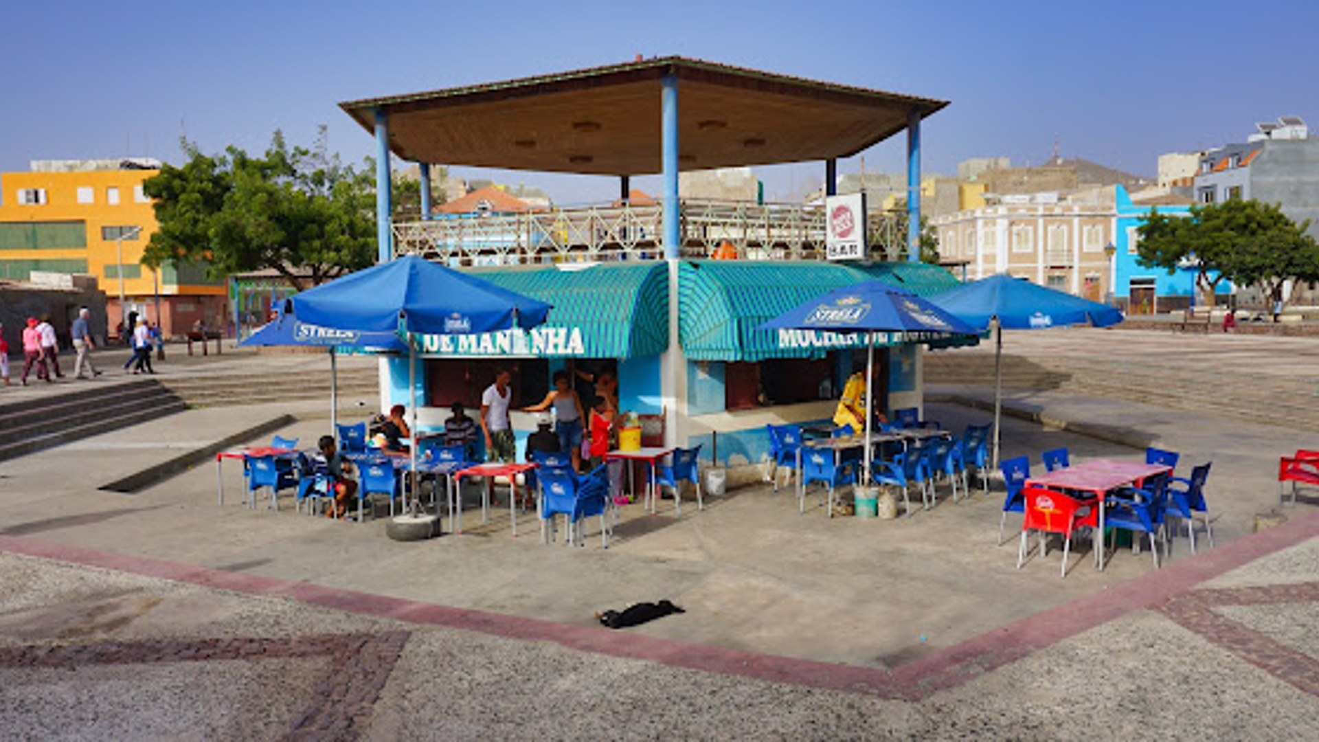 Outdoor food kiosk with blue awnings and tables in a paved square in Mindelo.